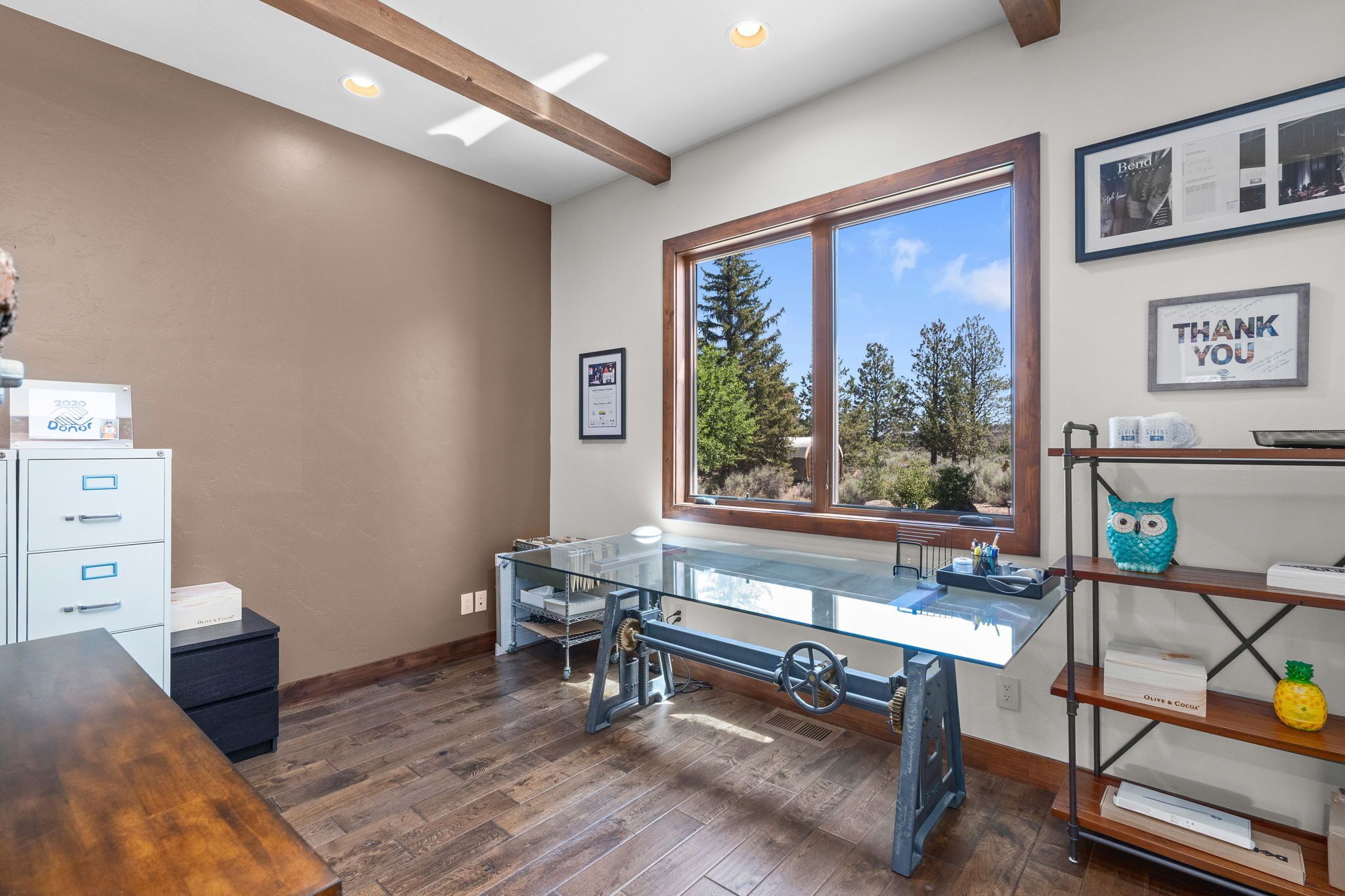 Una habitación con un ventanal grande, una mesa de vidrio, suelo de madera y una estantería metálica con un adorno de piña.