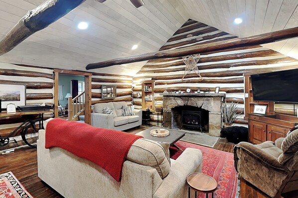 A log cabin interior with a fireplace, sofa, television, and a dining area.