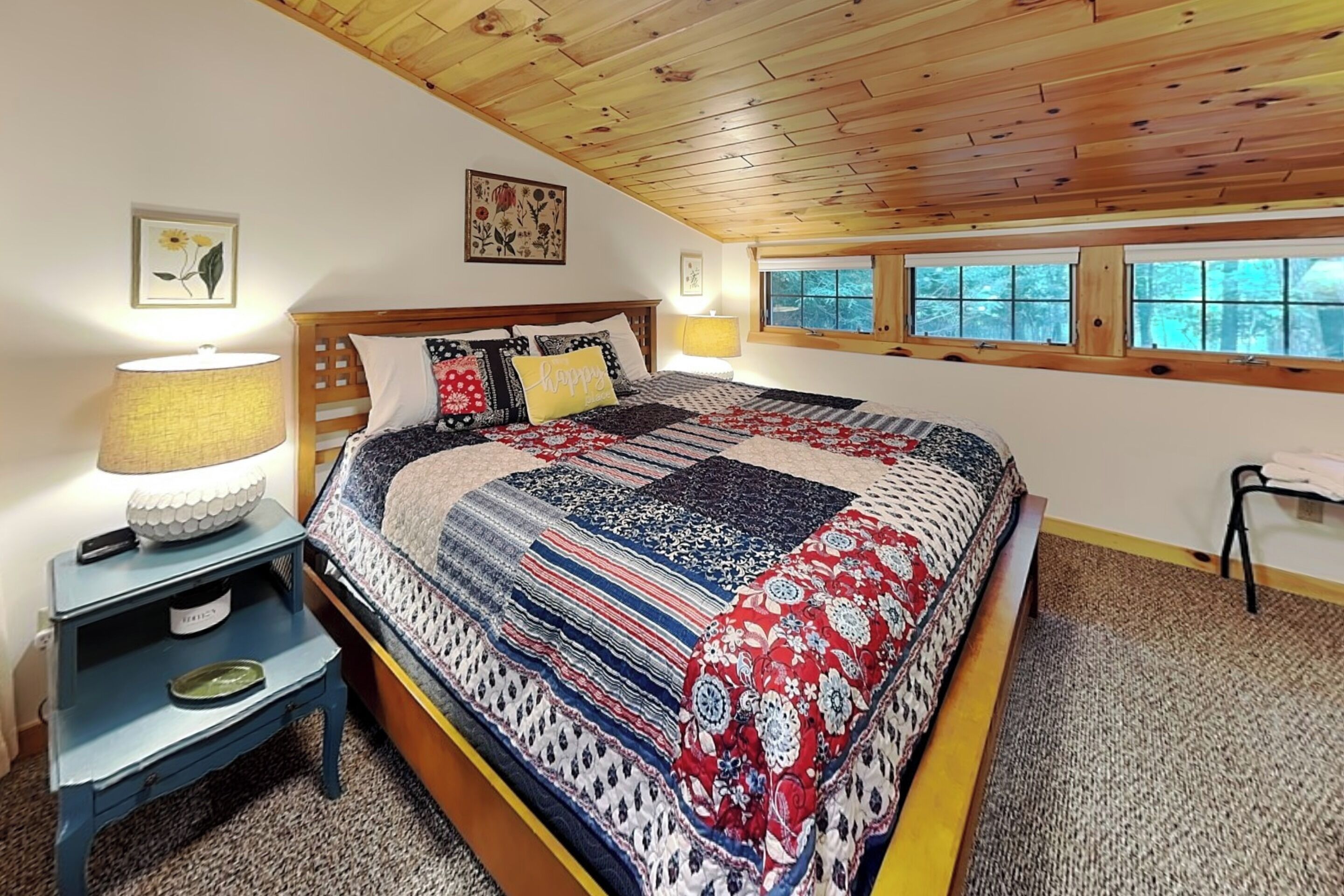 A bedroom with a wooden ceiling, a bed with a patterned quilt, bedside lamps, a nightstand, and a window with a view of trees.