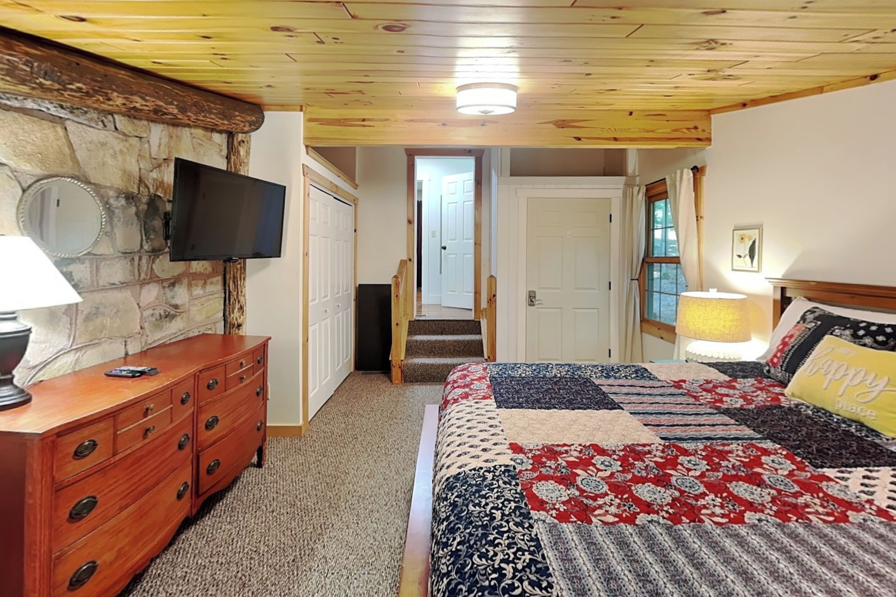A bedroom with a wooden ceiling, a stone wall, a flat-screen TV, a wooden dresser, a quilted bedspread, and a door leading to another room.