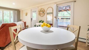 A dining area with a round table, chairs, and a bowl of lemons. There is a window with curtains, a door, and a view of greenery outside.