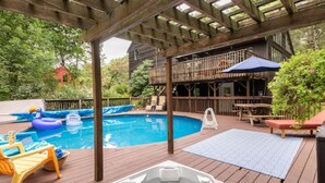 A wooden deck with a hot tub, a swimming pool, lounge chairs, and a covered area with a railing.