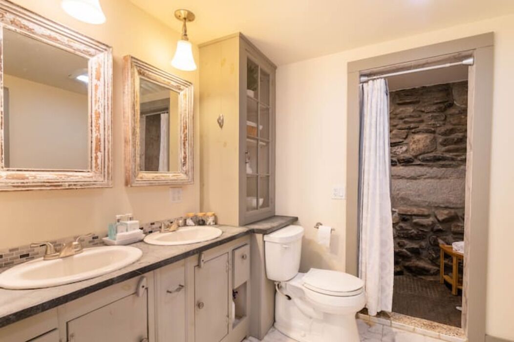 A bathroom with two sinks, a toilet, and a stone wall visible through an open door.