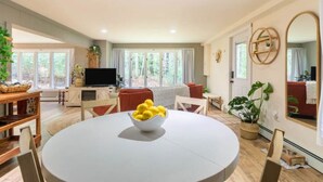 A spacious living room with a round dining table, a flat-screen TV, and a large window offering a view of greenery.
