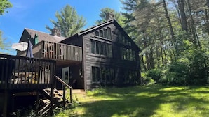 A two-story wooden cabin with a balcony, surrounded by a forested area.