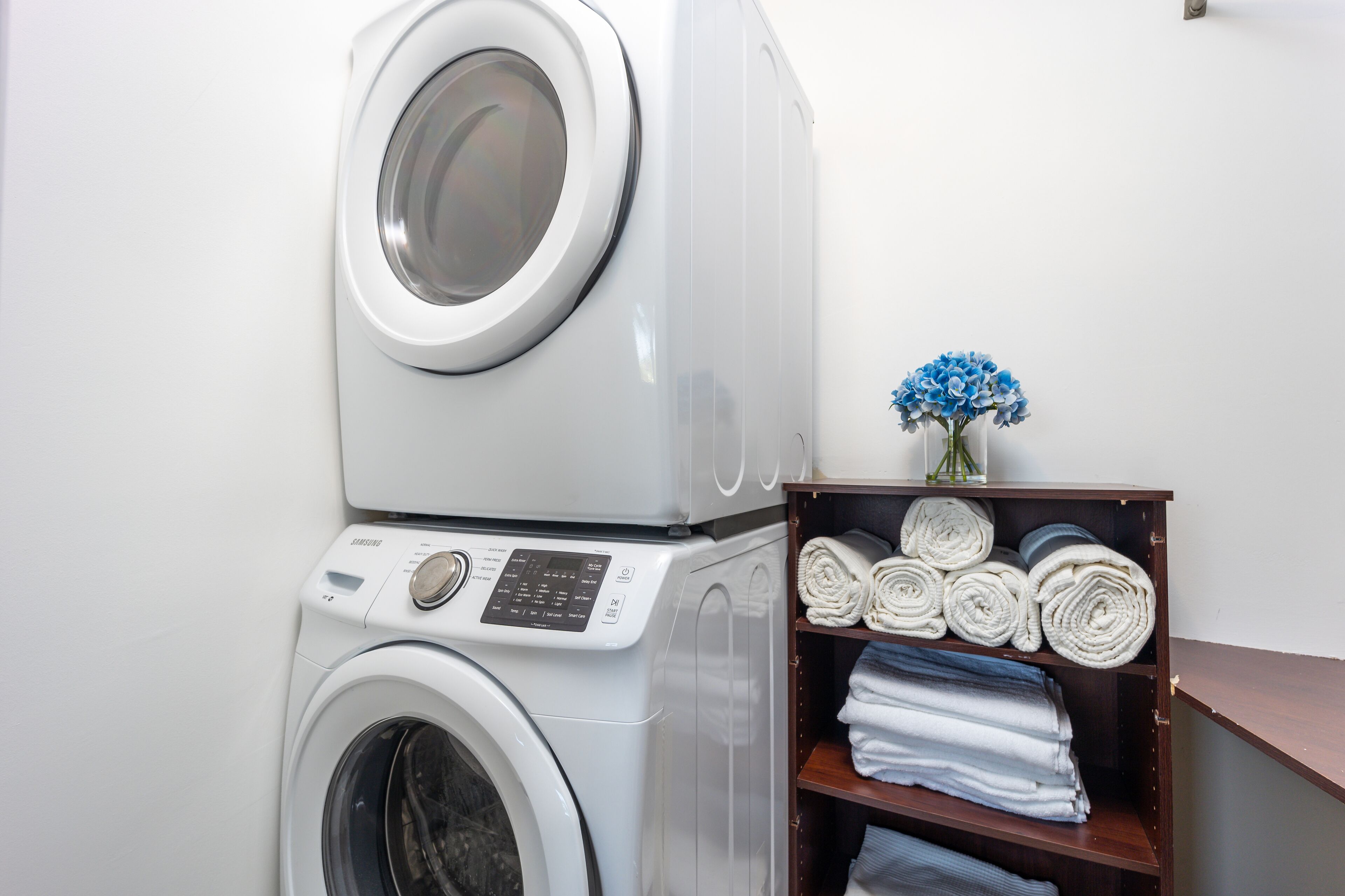 A stacked washer and dryer unit with a shelf of neatly rolled towels and a vase of flowers.