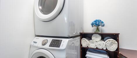 A stacked washer and dryer unit with a shelf of neatly rolled towels and a vase of flowers.