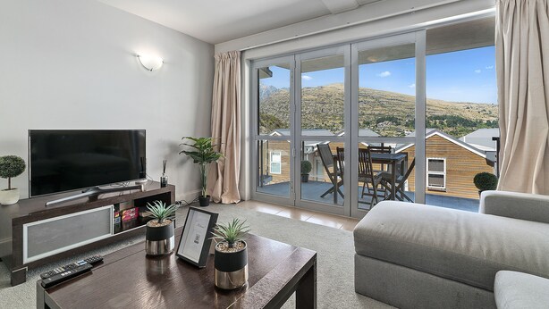 Een moderne woonkamer met een flatscreen televisie, een salontafel met decoratieve objecten, een grijze bank en een grote glazen schuifdeur die uitkomt op een balkon met een prachtig uitzicht.