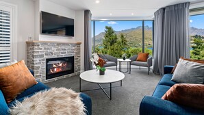 A modern living room with a stone fireplace, a flat-screen TV, and a view of mountains. - LOVELY 2 BEDROOM APARTMENT WITH INDOOR FIREPLACE (Queenstown)