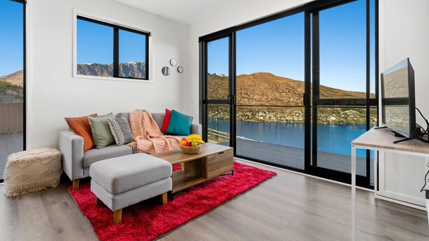 A modern living room with a large window overlooking a lake and mountains, a grey sofa with colorful cushions, a wooden coffee table, and a red rug. - A LUCKIE CHARM - 3-BEDROOM TOWNHOUSE WITH SPECTACULAR VIEW (Queenstown Hill)