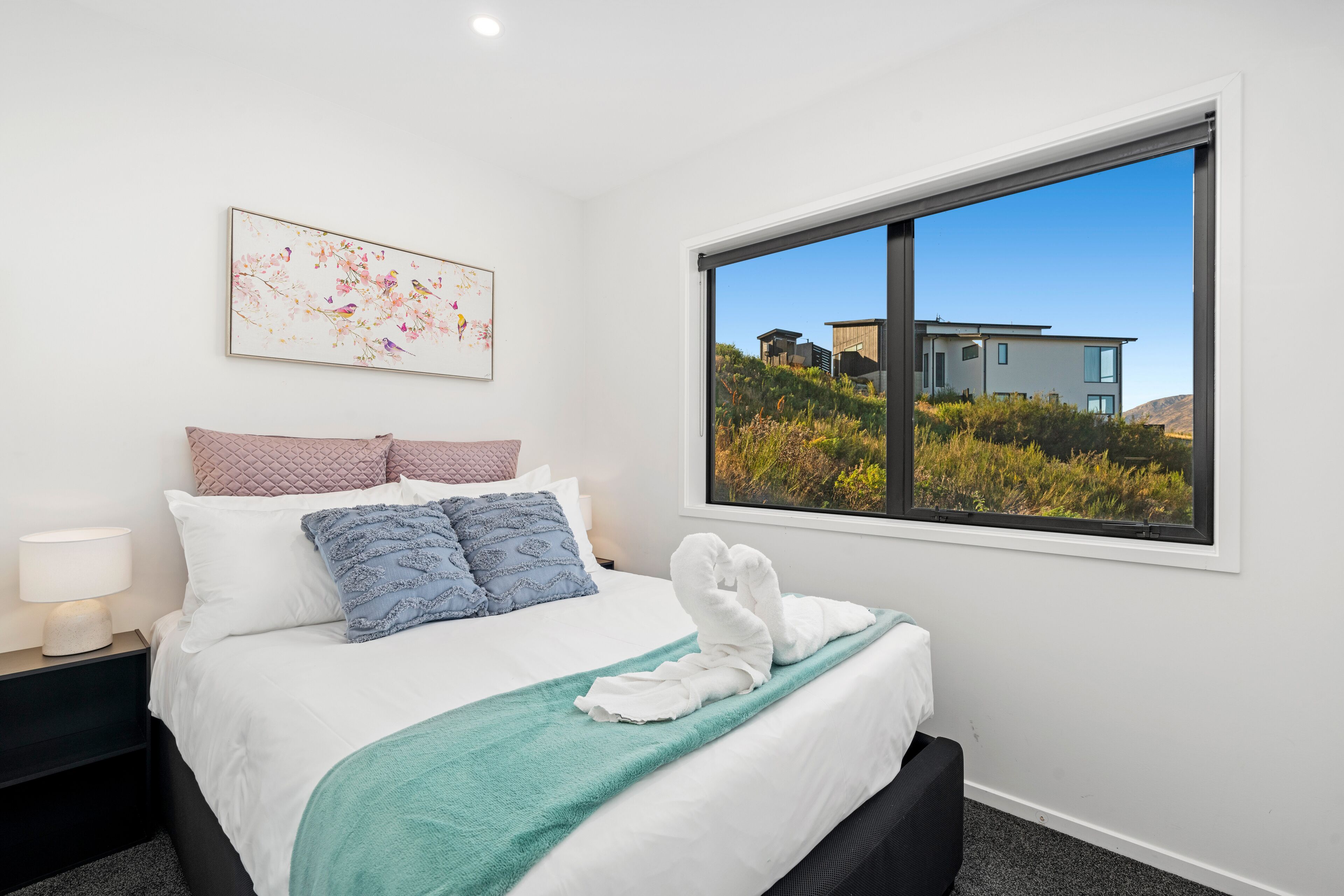A neatly made bed with a teal throw blanket, a white towel, and a decorative pillow. A framed artwork is on the wall above the bed, and a window offers a view of a modern house and greenery.