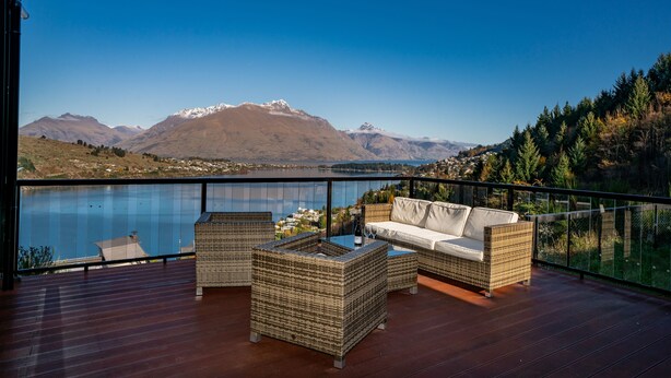 A balcony with wicker furniture, a glass table, and a view of mountains and a lake.