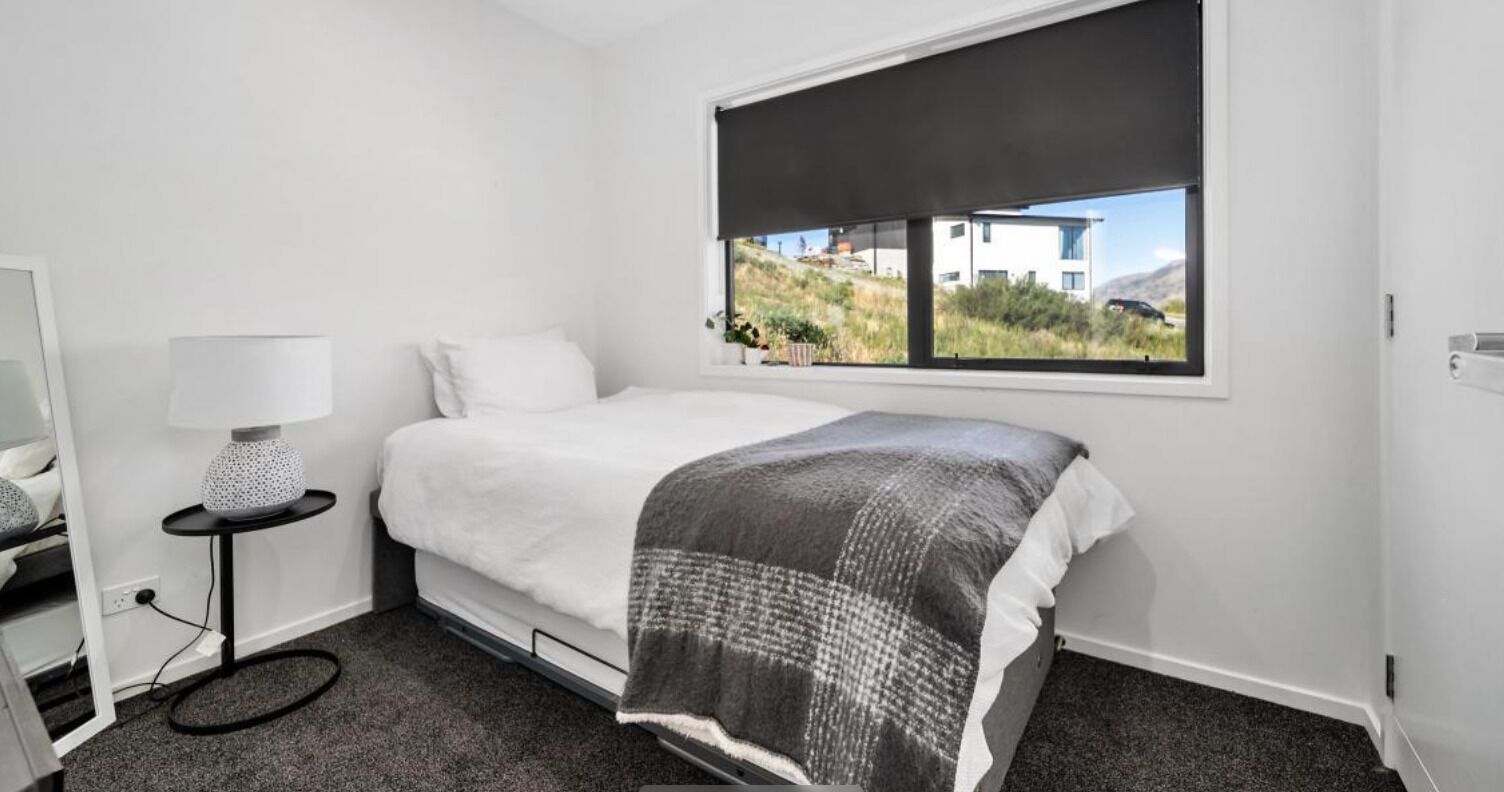 A bedroom with a bed, a bedside table with a lamp, a window with a view of buildings and hills, and a carpeted floor.