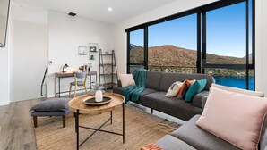A modern living room with a large window, a gray sofa, a wooden coffee table, and a desk with a chair. - A LUCKIE CHARM - 8 BEDROOM TOWNHOUSE (Queenstown Hill)
