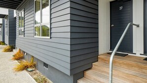 A modern house with a dark gray siding exterior, a black front door with the number 27, and a wooden front porch with a metal handrail. - 2-BEDROOM APARTMENT - BEECHWOOD (Queenstown)