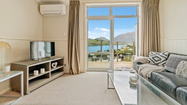 A modern living room with a television, a sofa, a coffee table, and a view of a mountainous landscape through a large sliding glass door. - 2-BEDROOM APARTMENT - BEECHWOOD (Queenstown)