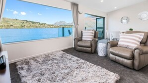 Uma sala de estar moderna com uma grande janela que oferece vista para um lago e montanhas. Há dois sofás de couro com almofadas listradas e um tapete felpudo no chão.
