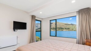 Quarto com cama grande, TV de tela plana na parede e janela com vista panorâmica para um lago e montanhas.