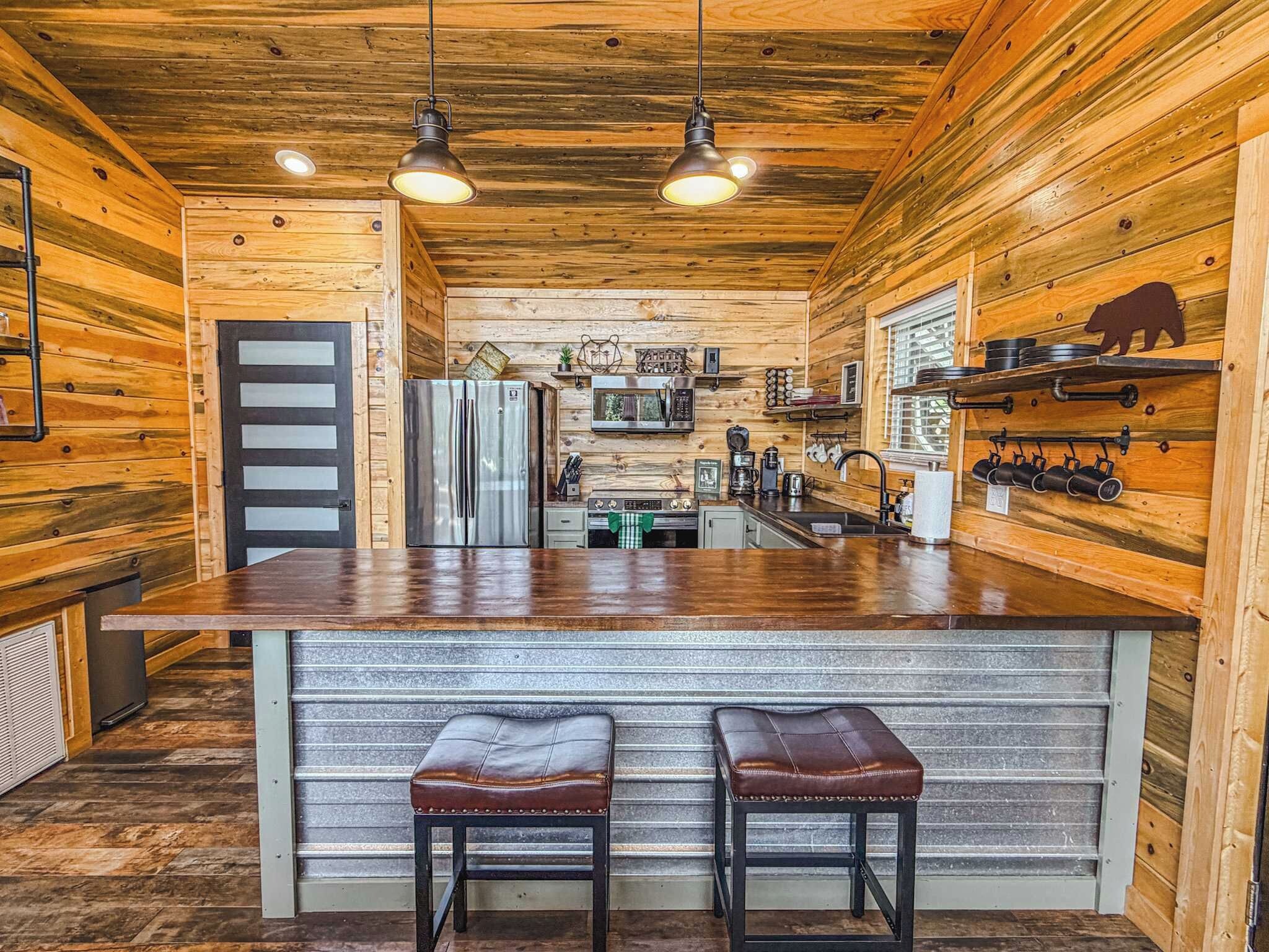 Una cocina con paredes de madera, un refrigerador de acero inoxidable, un microondas y un lavamanos. Hay dos taburetes en la isla.