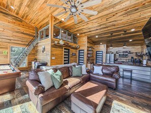 A spacious cabin interior with a high ceiling, a central seating area with a sofa and ottoman, a kitchen area, and a staircase leading to an upper level.