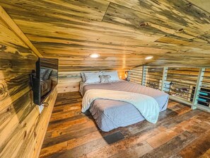 Een houten plafond, een bed met een deken, een aan de muur bevestigde televisie en een houten vloer.
