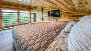 Ein Schlafzimmer mit Holzboden, einem Bett mit gemustertem Überwurf, einem an der Wand befestigten Fernseher und großen Fenstern.