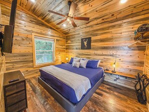 A wooden cabin bedroom with a bed, ceiling fan, window, and wall-mounted light fixture.