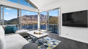 A modern living room with a large TV, a sofa, a coffee table with wine glasses, and a view of mountains and a lake through large windows. - MODERN ALPINE LIVING SPECTACULAR VIEW (Queenstown)