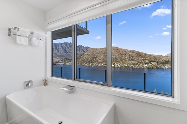 A bathroom with a large bathtub, a towel rack with two towels, and a window overlooking a lake and mountains. - MODERN ALPINE LIVING SPECTACULAR VIEW (Queenstown)