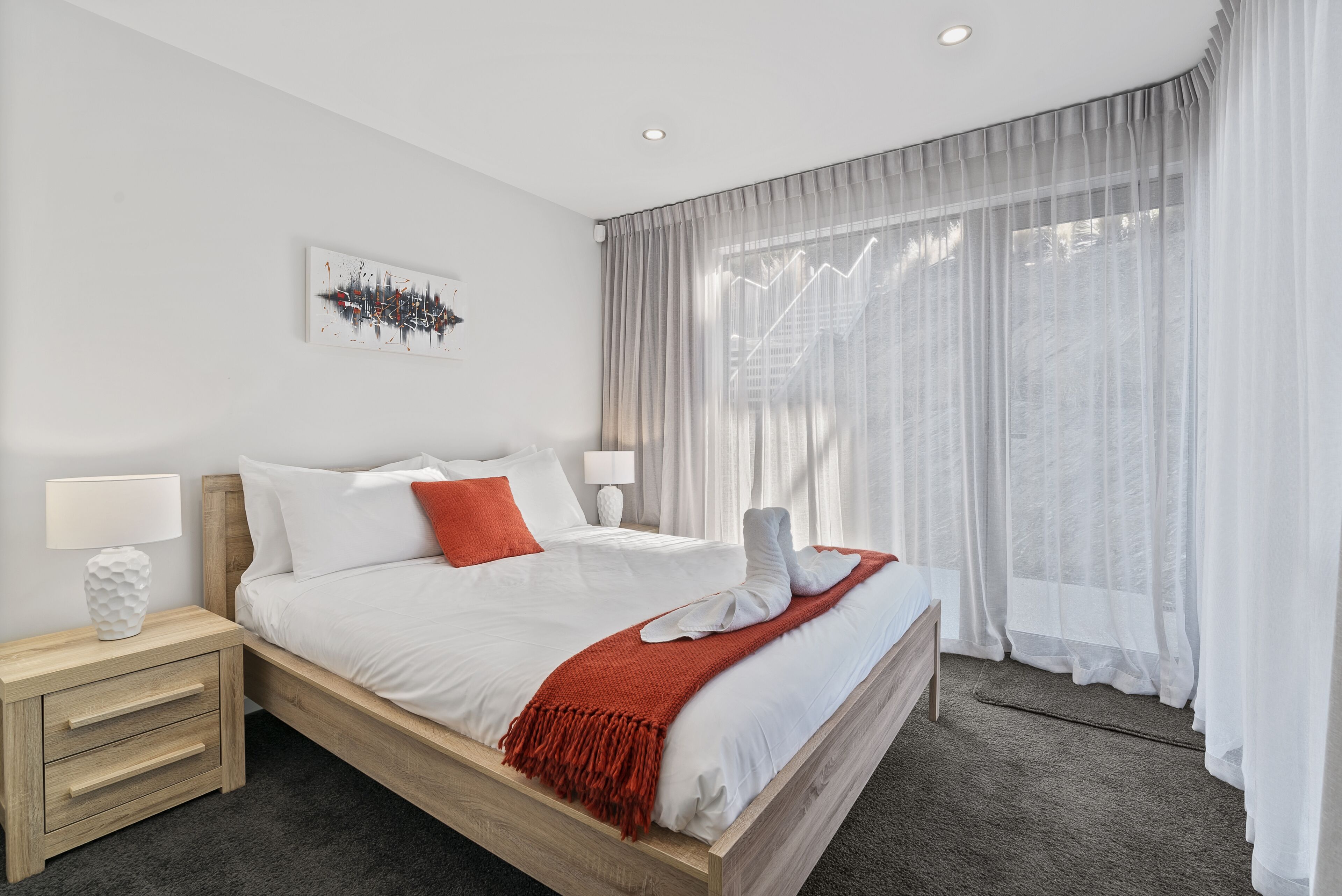 A modern bedroom with a wooden bed, white bedding, a red throw blanket, and sheer curtains.