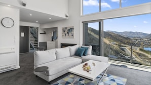 A modern living room with a large window, a sofa, a coffee table, and a staircase leading to an upper level. - MODERN ALPINE LIVING SPECTACULAR VIEW (Queenstown)