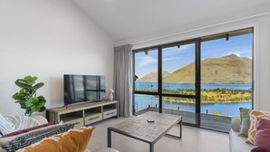 Un salon moderne avec une grande télévision, une table basse en bois et un canapé. Il est doté d’une grande porte-fenêtre coulissante offrant une vue panoramique sur les montagnes et le lac.