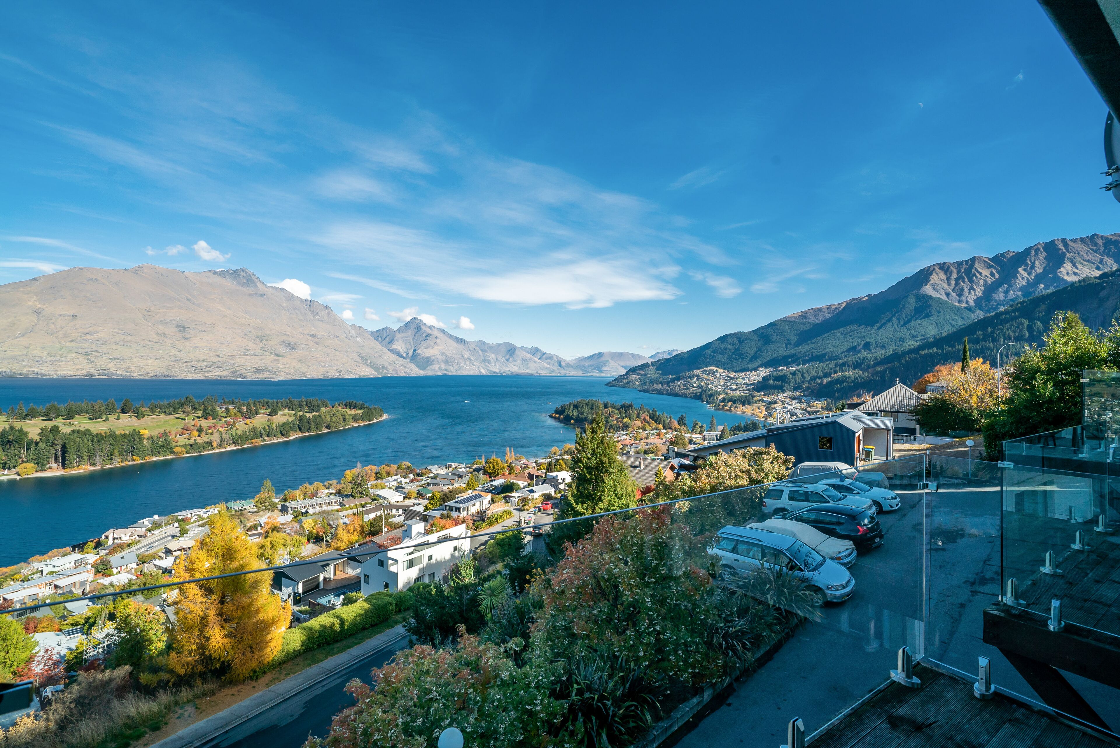 A scenic view of a lake with mountains in the background, a parking area with cars, and a building with a glass facade.