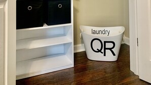 En vasketøjskurv med påskriften 'laundry QR' og et hvidt opbevaringsskab med hylder og mulighed for at hænge tøj.