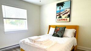 Ein ordentlich bezogenes Bett mit weißer Bettdecke und gefalteten Handtüchern, ein hölzerner Kopfteil und ein Poster mit einer Strandansicht, auf dem 