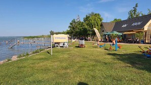 Une zone au bord du lac comprenant un espace de baignade, une terrasse enherbée avec des meubles d’extérieur et un bâtiment sur lequel est apposée une enseigne indiquant « VERLEIH ».
