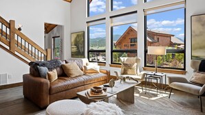 Ein geräumiges Wohnzimmer mit braunem Ledersofa, einem Couchtisch und Blick auf eine Holzhütte durch große Fenster.