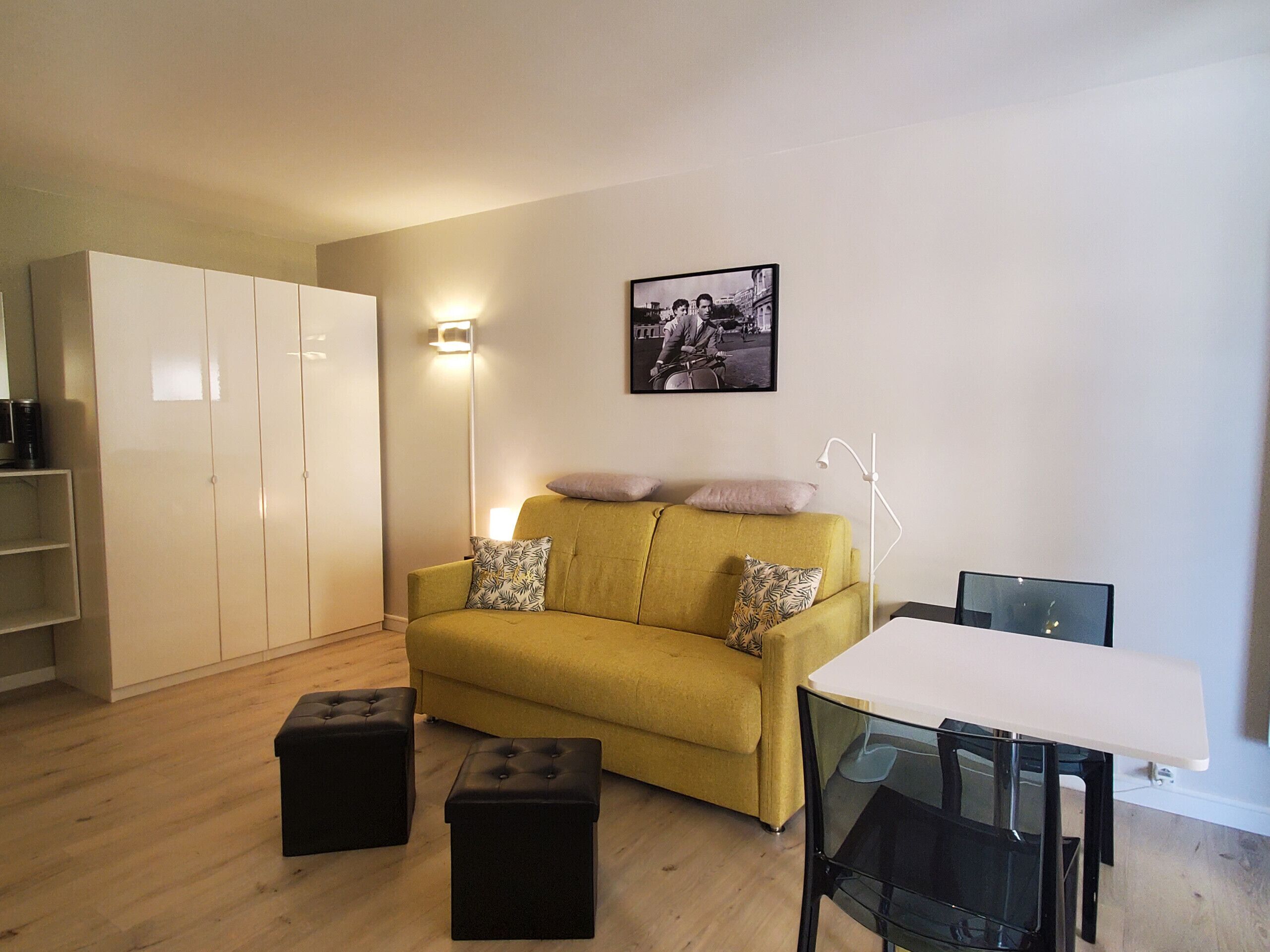 Ein Wohnzimmer mit gelbem Sofa, zwei schwarzen Hocker, einem weißen Tisch und einem schwarzen Sessel. Es gibt einen weißen Kleiderschrank, eine Stehlampe und ein gerahmtes Bild an der Wand.