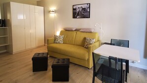 Una sala de estar con un sofá amarillo, dos otomanes negros, una mesa blanca y una silla negra. Se observa un armario blanco, una lámpara de pie y un cuadro enmarcado en la pared.