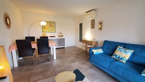 Een woonkamer met een blauwe bank, een eethoek met een tafel en stoelen, en een keuken op de achtergrond.