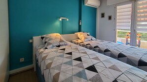 Slaapkamer met twee bedden, teal muren, een aan het plafond gemonteerde airconditioner, een bedlampje en een raam met jaloezieën.