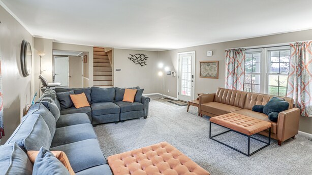 A spacious living room with a sectional sofa, a loveseat, and an ottoman. There is a staircase leading to another level, a large mirror, and a window with curtains.