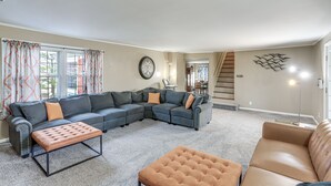 A living room with a sectional sofa, ottomans, a clock, and a staircase leading to another room.