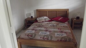 Una cama con cabecera de madera, un almohilarrojo y una manta con la palabra 'Voyage' escrita. Hay dos mesitas de noche con lámparas y un estante pequeño.