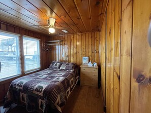 Habitación con paneles de madera, una cama, una mesita de noche, una lámpara y un ventilador de techo.