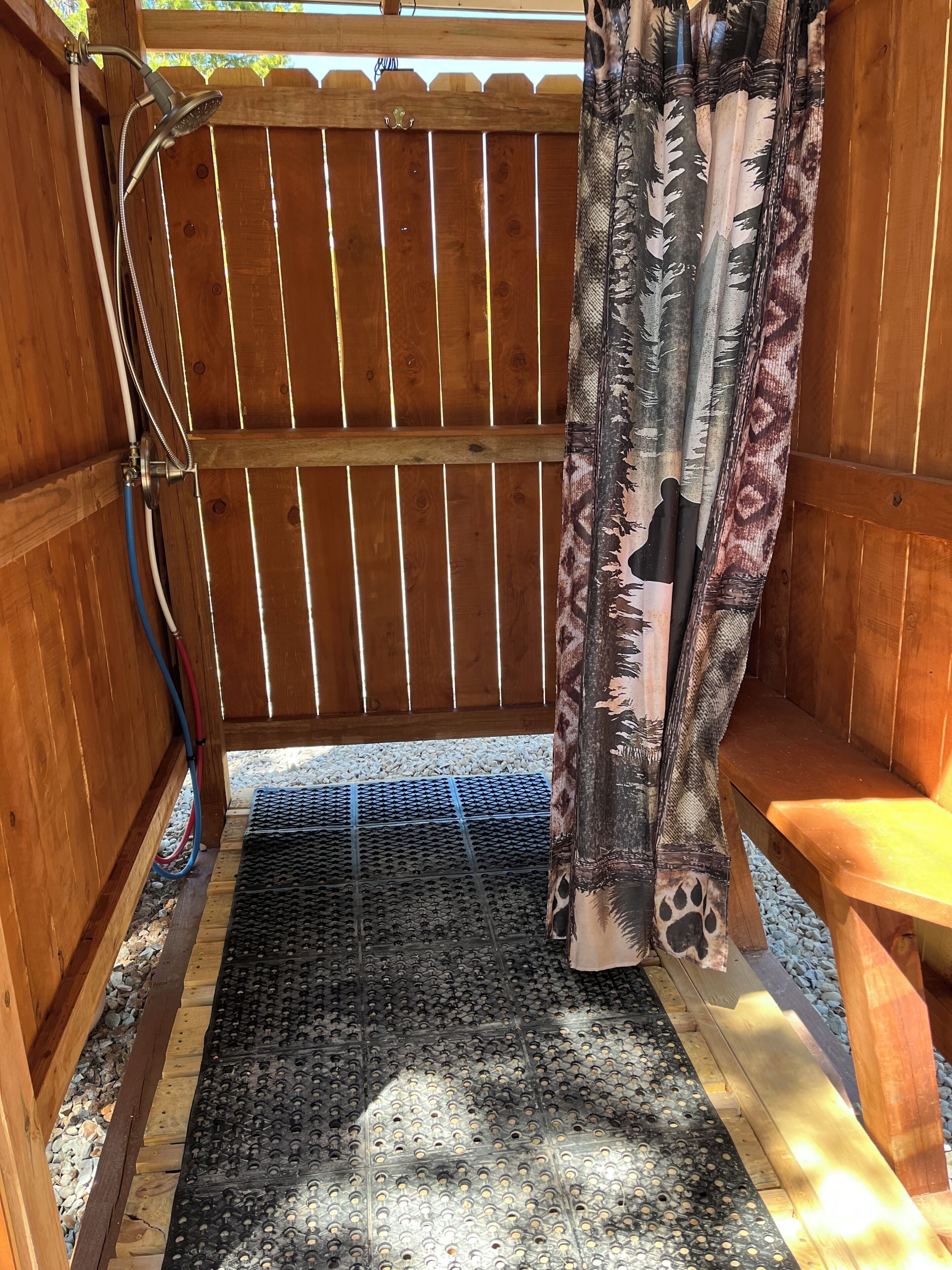A wooden shower enclosure with a metal showerhead, a black textured mat, and a wooden bench.