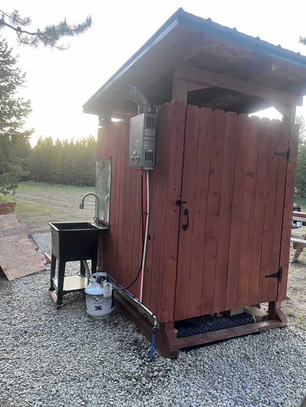 Un cobertizo de madera con un calentador de agua metálico, un lavamanos y un tanque de propano.