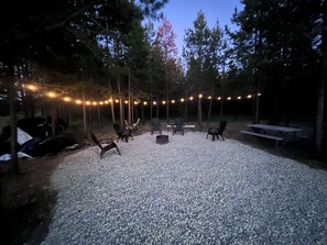 Un espacio al aire libre con sillas y una chimenea, rodeado de árboles y luces colgantes.