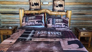 Een eenvoudige slaapkamer in een houten blokhut met een houten bed, meerdere kussens met dieren erop en een quilt met de tekst 'ADVENTURE'.