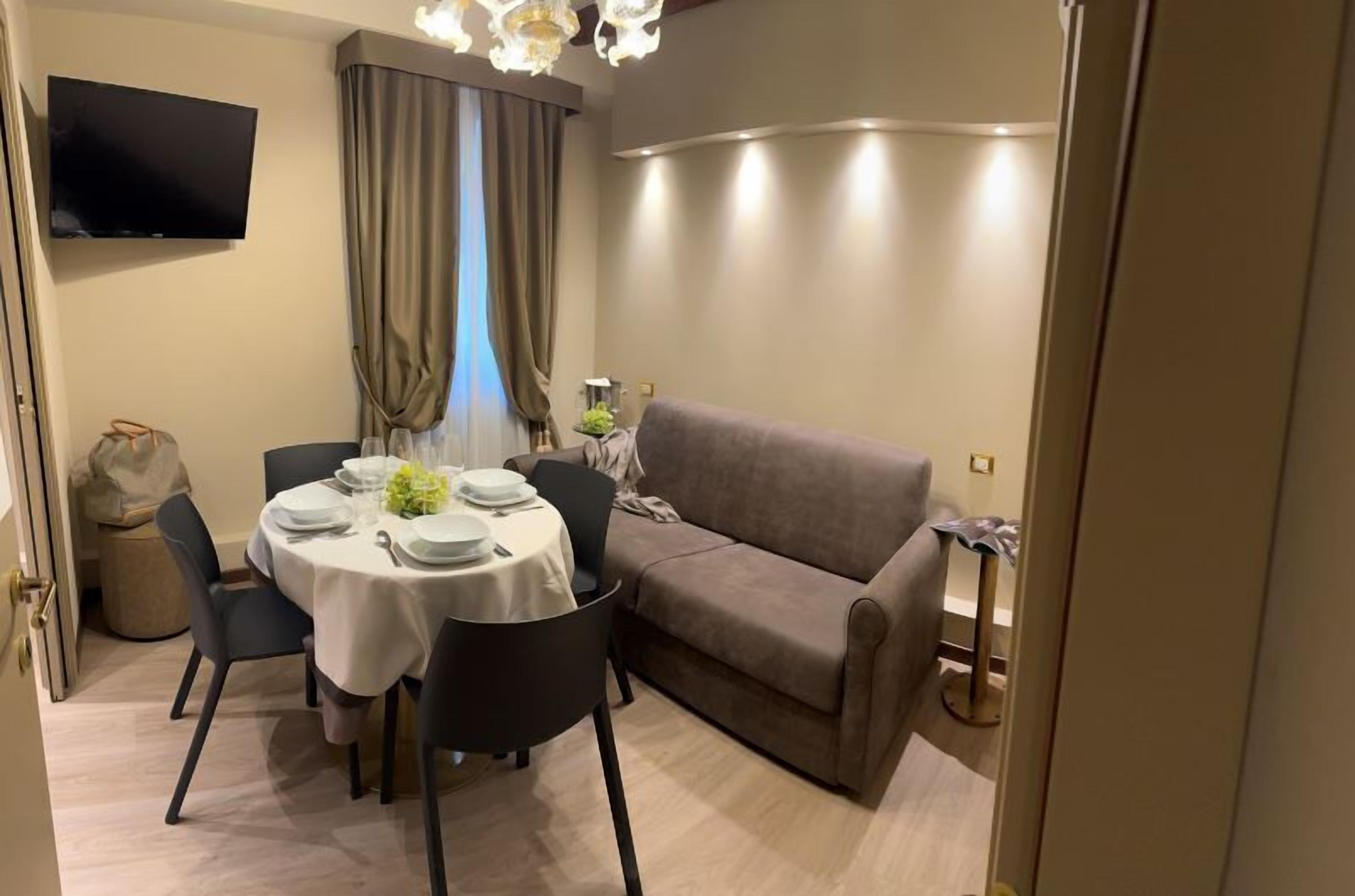 A dining area with a round table set for four, a sofa, a television mounted on the wall, and a window with curtains.